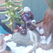 A crimson-armored space alien sits on a stone next to its helmet amidst white pebbles and succulents.