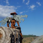 A brown Bionicle with robotic wings perched on a wooden fencepost looking over a field on a clear day.