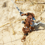 Top-down view of a brown Bionicle holding robotic wings to climb up a steep sand cliff.