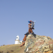 Low angle shot of a brown Bionicle with robotic wings perched on a rock with a lighthouse on a green hill in the background.
